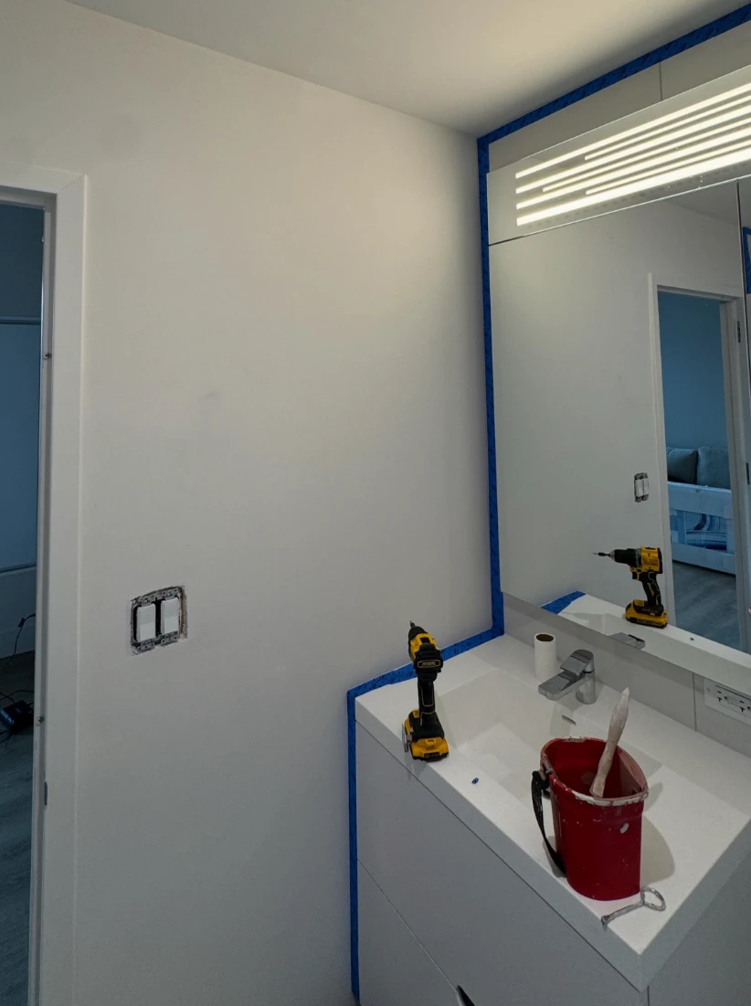 Apartment bathroom before painting, with plain white walls and ceiling and blue painter's tape masking the mirror and vanity edge