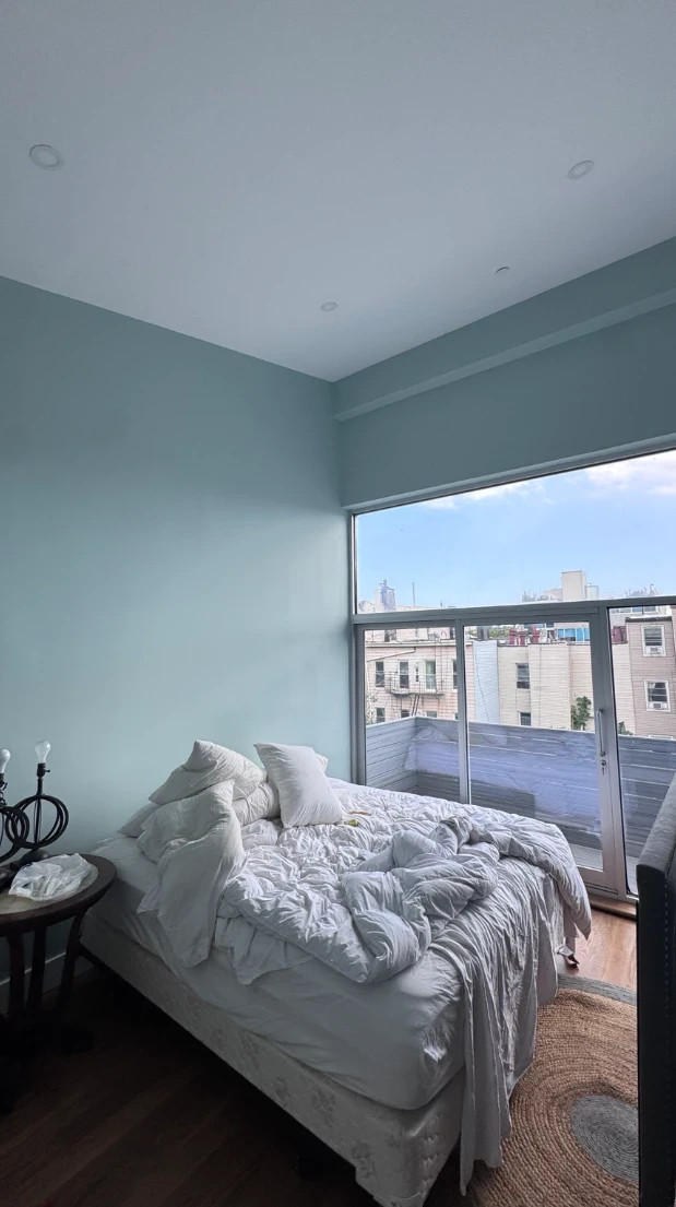 The same bedroom after painting, with soft aqua walls, a large corner window with a balcony view, and a bed with white bedding
