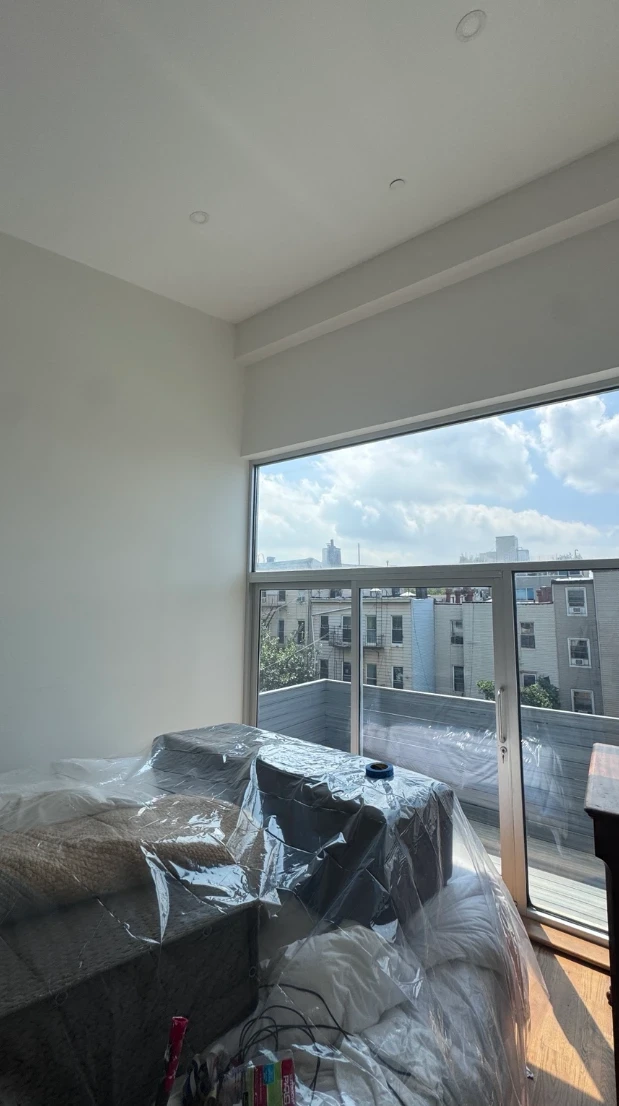 Bedroom before painting, with plain white walls, a large corner window with a balcony door, and furniture covered in plastic sheeting
