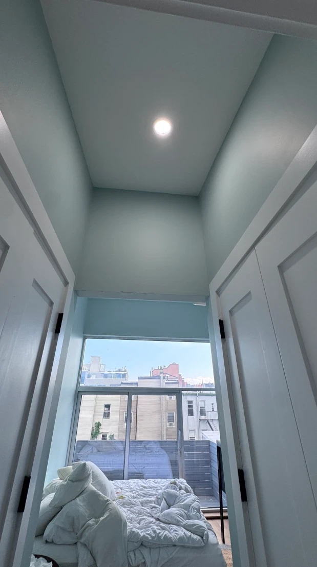 View through a doorway into a soft aqua bedroom alcove with a recessed ceiling light, leading to the bedroom window and balcony