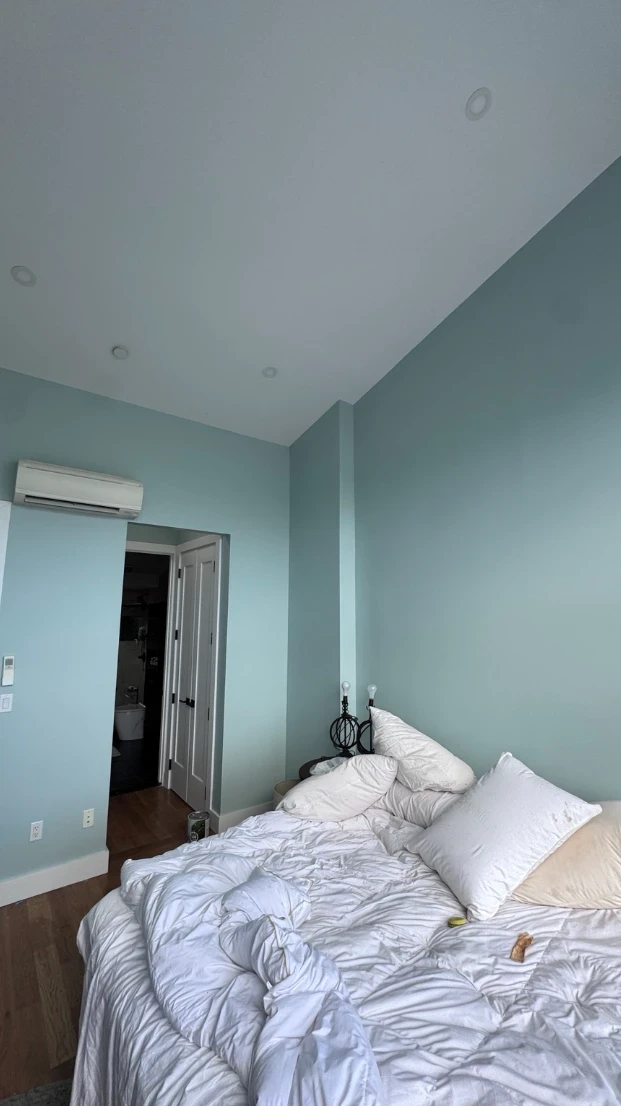 Bedroom with soft aqua walls, a wall-mounted air conditioner, a doorway to the bathroom and a bed with white bedding