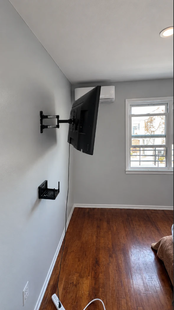 Apartment bedroom wall painted light gray with a wall-mounted TV on a swivel arm, a mini-split air conditioner, a white-framed window and white baseboards above dark hardwood floors