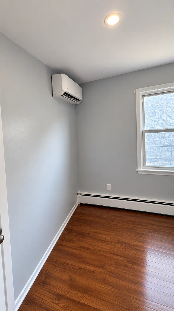 Apartment bedroom with smooth light gray walls, a white-framed window, a wall-mounted air conditioner, a white baseboard heater and warm hardwood floors