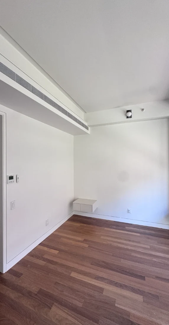 Apartment bedroom wall painted smooth white with a long linear ceiling vent, white baseboards, a small white wall-mounted shelf and hardwood floors