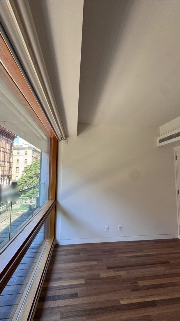 Corner of an apartment bedroom with smooth white walls and ceiling and a tall wood-framed window with a view of the street, above warm hardwood floors