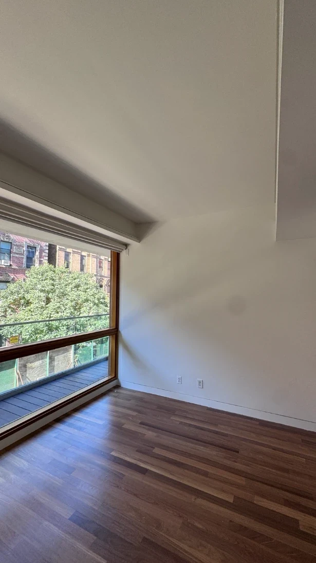 Apartment bedroom with smooth white walls and ceiling, a large wood-framed window with a roller shade looking out to trees and brick buildings, and warm hardwood floors