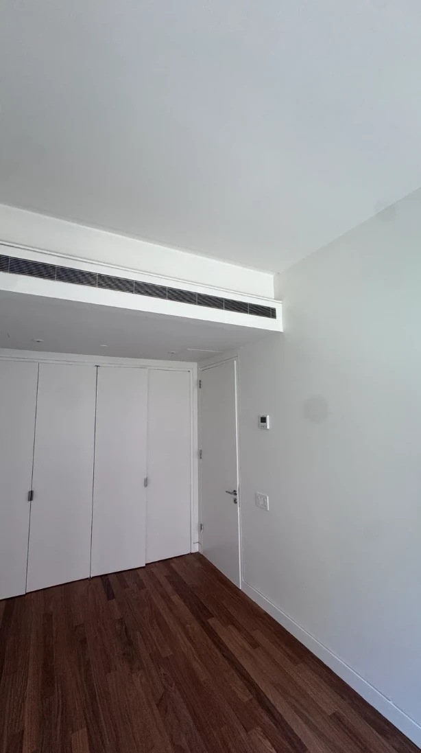 Apartment bedroom with smooth white walls and ceiling, a long linear ceiling vent, white closet doors, a flat white door and hardwood floors
