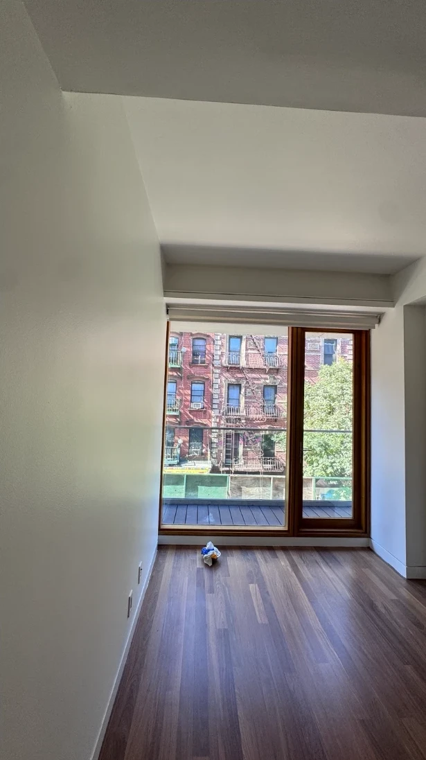 Apartment bedroom with smooth white walls and ceiling, a wood-framed floor-to-ceiling window with a roller shade looking out to a brick building, and warm hardwood floors