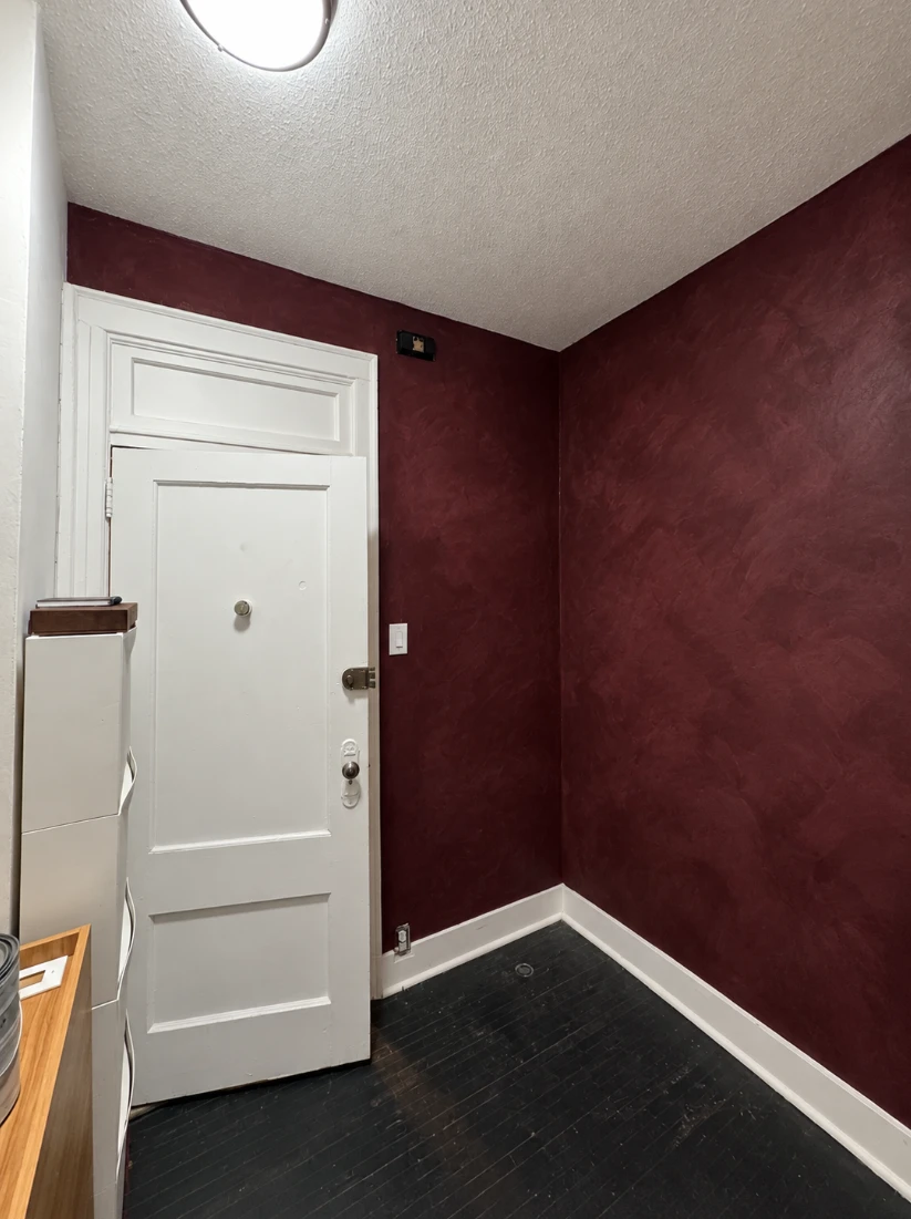 Apartment room with deep burgundy lime wash walls, a white paneled door with a transom above it, white baseboards and a dark wood floor