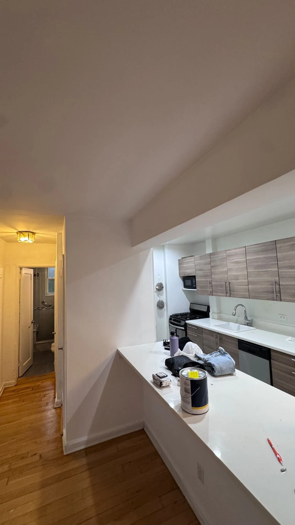 Open apartment kitchen and hallway with fresh white walls and ceiling, a white countertop peninsula, wood-look cabinets and hardwood floors