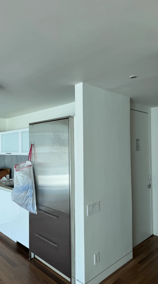 Apartment kitchen and entry area with smooth white walls and ceiling, a stainless steel refrigerator, a white door and dark hardwood floors