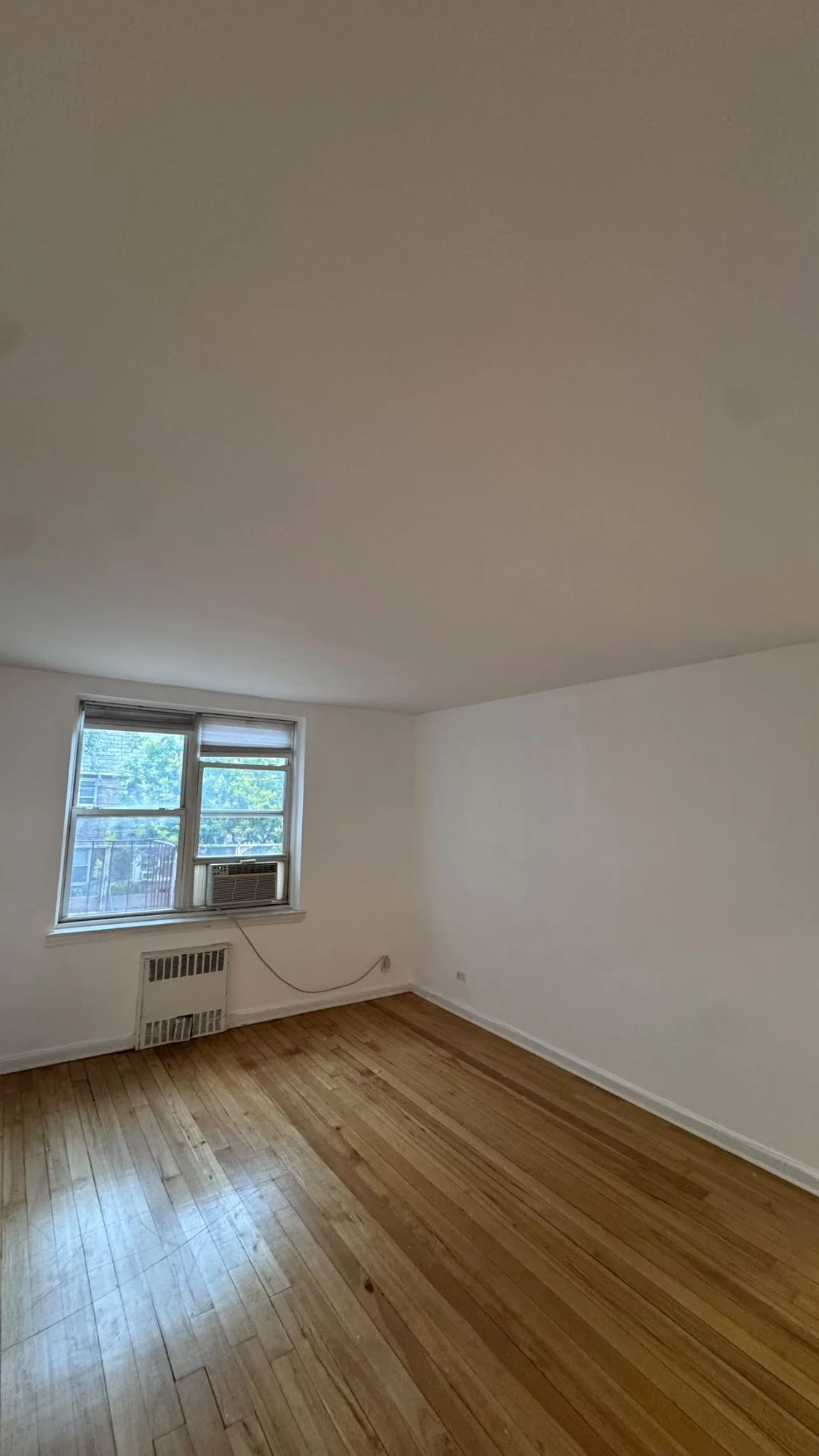 Empty apartment room with fresh white walls and ceiling, a window with a radiator cover below it, and hardwood floors