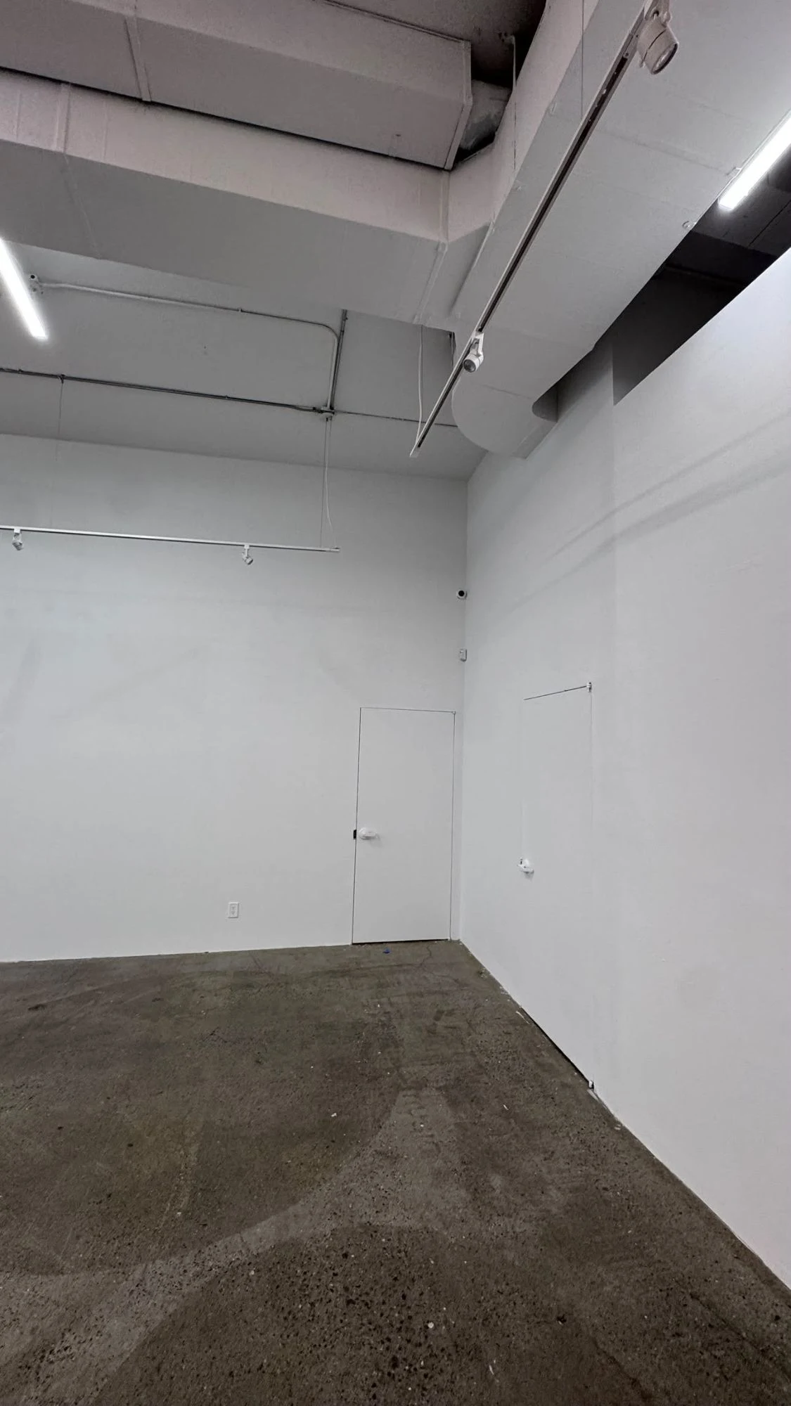 Corner of a commercial space with tall smooth white walls, two white flush doors, track lighting and white ducts above a concrete floor